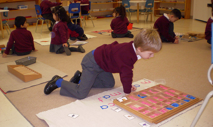 Create a realistic image of a diverse Montessori classroom with children aged 3-6 engaged in various activities. A white female child works with colorful wooden blocks, a black male child tends to a small plant, and an Asian female child uses a sandpaper letter. The room features low shelves with Montessori materials, a cozy reading nook, and large windows letting in natural light. A banner on the wall reads "Nurturing Independence and Creativity".